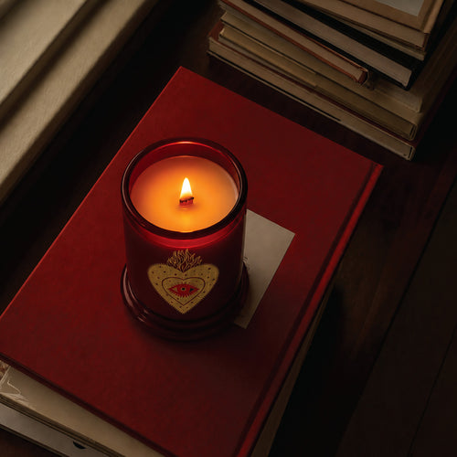 A lit red glass candle jar with a heart emblem sits on a red book on a dark wooden surface, with books stacked in the background.