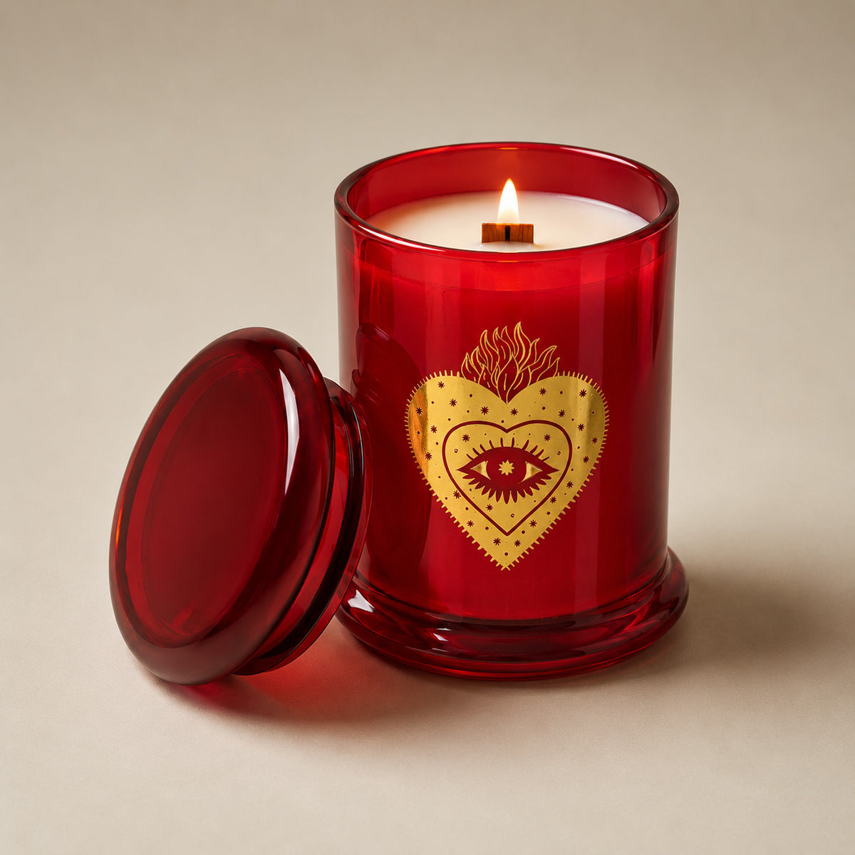 A red glass jar candle with a white wax fill and a lit wick, featuring a gold sacred heart design with an eye motif on the front. The matching red glass lid rests beside the candle on a neutral beige background.